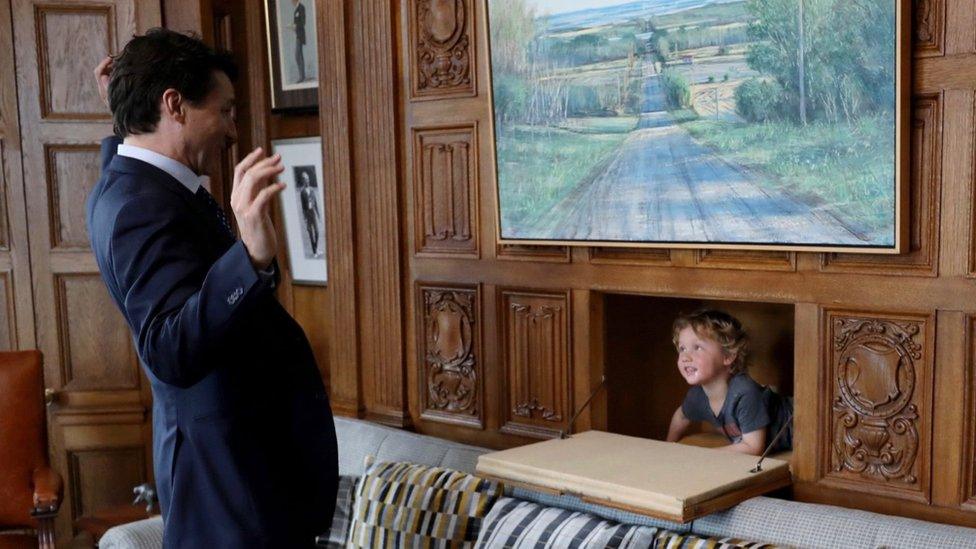 Justin's son emerges from a hidden compartment in the wall through a door in the wood panelling, while a smiling Justin throws his arms up in the air in mock surprise