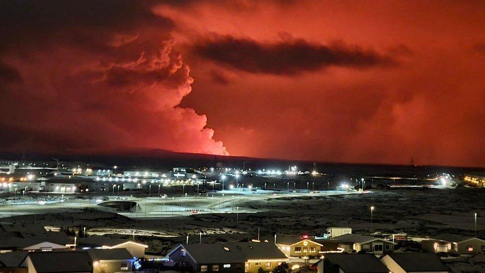 Houses in the village of Hafnarfjordur is seen as smoke is billowing in the distance as the lava colour the night sky orange from an volcanic eruption on the Reykjanes peninsula, western Iceland on December 18, 2023.