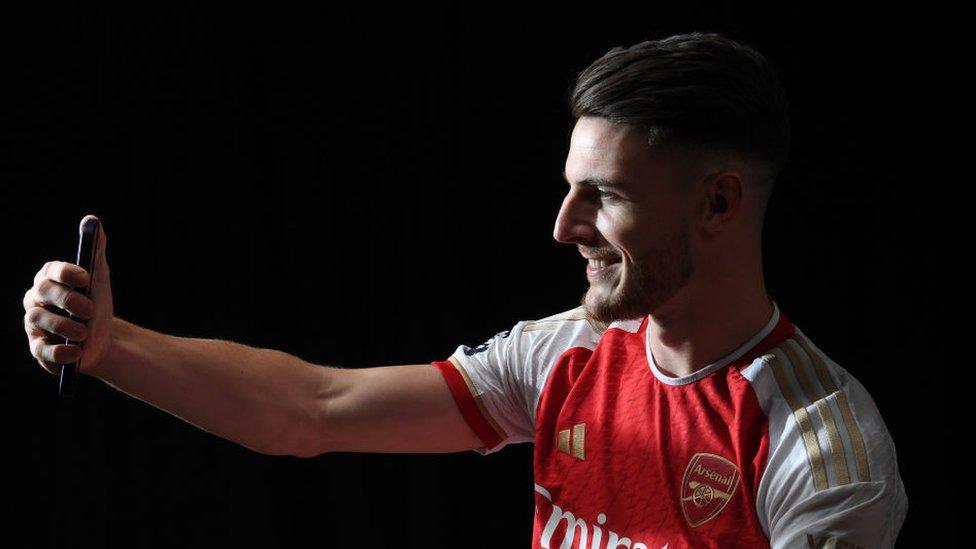 Footballer Declan Rice sitting side on and holding a mobile phone in his right hand with his right arm outstretched as if he is taken a selfie picture or video of himself. He is smiling and has his black hair slicked back over his head. He is wearing the Arsenal home kit which is mainly red with white sleeves. The background behind him is totally black.