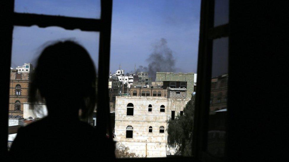 The silhouette of a girl looking out a window as plumes of smoke.