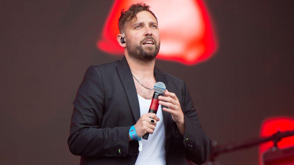 You Me At Six's Josh Franceschi performing at Reading festival - he is wearing a black blazer, a white t-shirt and is holding a microphone