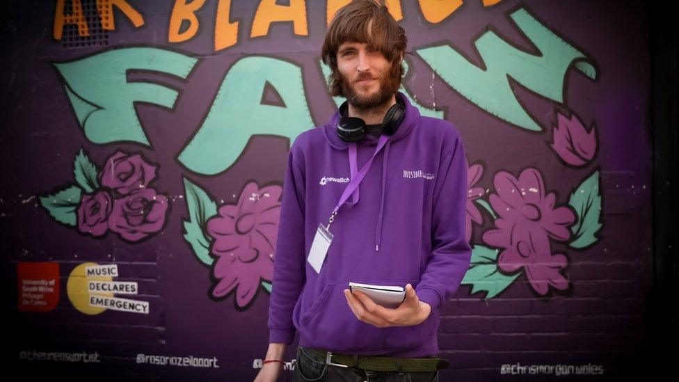 Johnny Giles in a hoodie with long bangs and a beard standing in front of a mural, holding a note pad