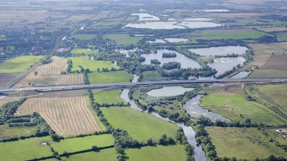 An aerial view of the new A14 after it opened. It shows the road raised up above a patchwork of ploughed and grassy fields. In the middle is a network of tree-lined ponds and a river.