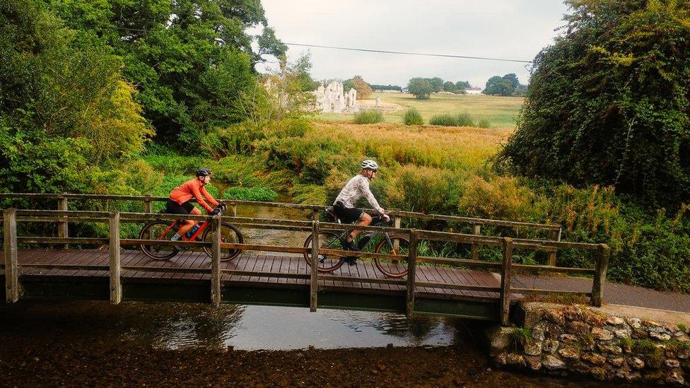 A man and a woman riding over a bridge spanning a ford in Castle Acre with the ruins of the priory in the background, on the Rebellion Way, Norfolk