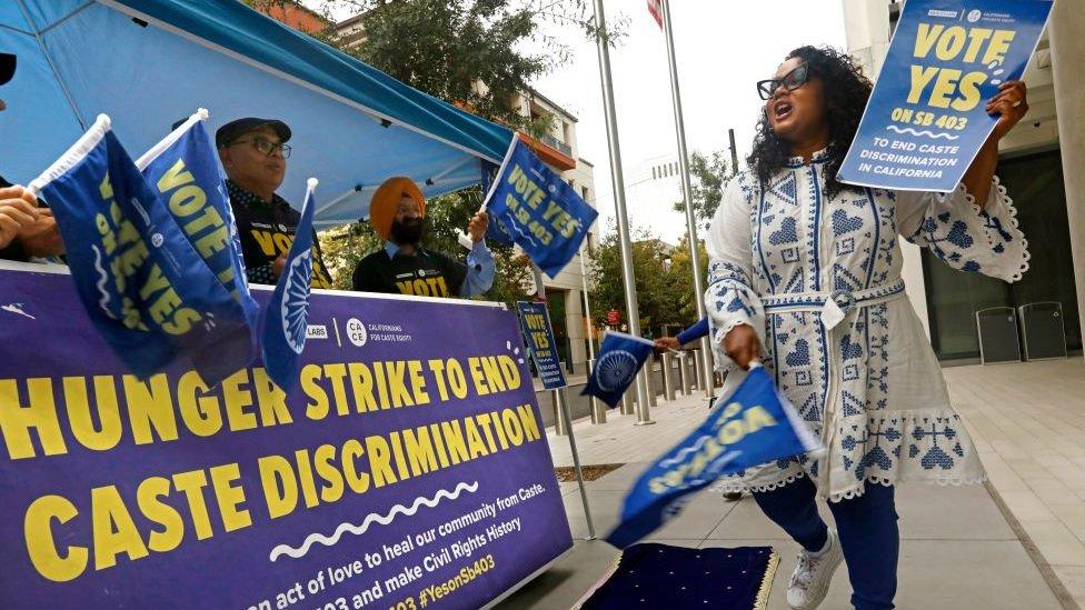 Woman in blue and white dress holding sign that says "Vote yes"in front of blue and yellow banner announcing hunger strike.