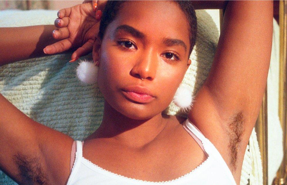 A young black woman poses with her arms above her head, showing her armpit hair