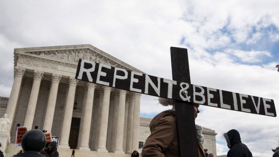 Protester holds sign shaped like a cross reading: 'Repent and Believe'