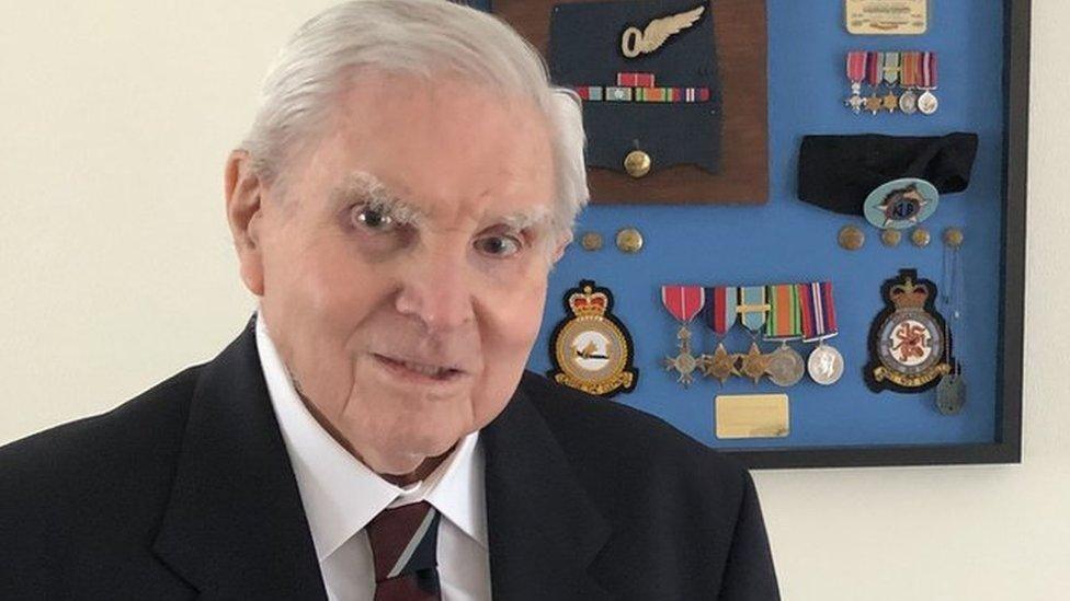 Stanley Booker posing for the camera. He has grey hair and is wearing a white shirt and RAF tie with a black jacket. On the wall behind him is a frame containing his medals, uniform buttons and badges.