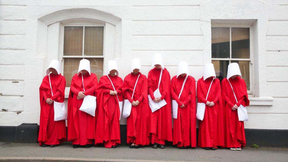 A group of handmaids in red cloaks and white hats with their heads bowed