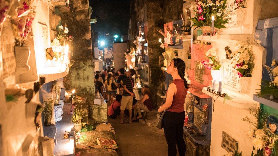 A woman stares at a group of colourful gravestones, behind her people are gathered at night in a well lit cemetery.