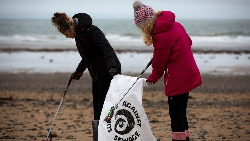 Two young people picking litter on the beach, bag reads "surfers against sewage"