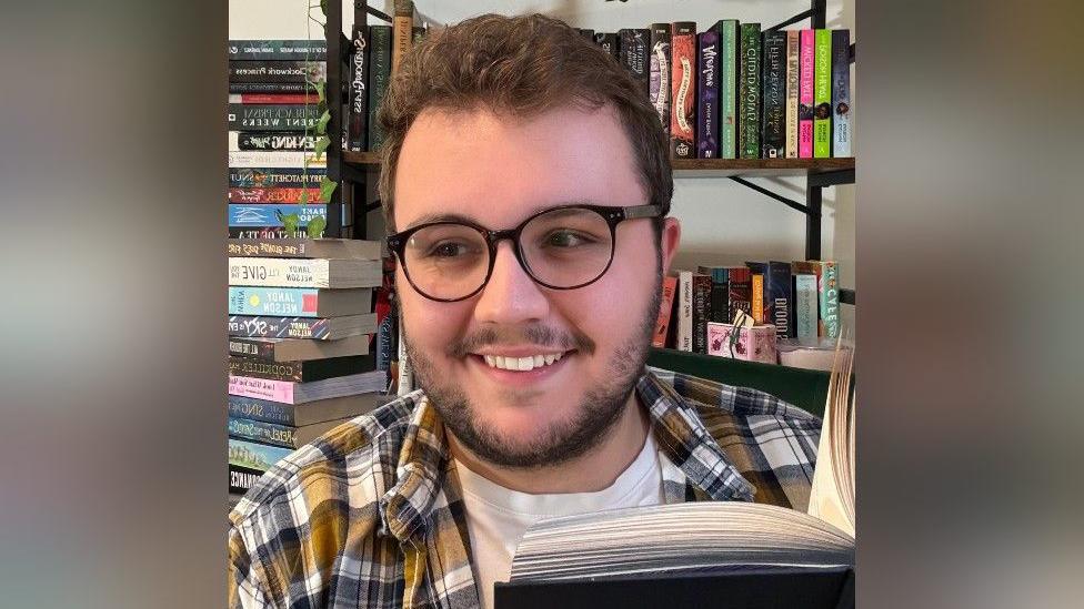 Bill Wood has brown hair and brown facial hair. He is wearing black glasses and a yellow, white and black shirt. He is holding a black book that is open in front of him.