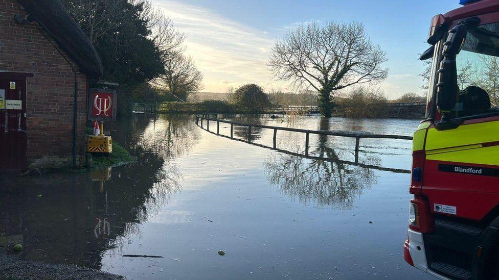 Fire engine stopped on edge of flooded road with building on left and wooden fence on right.