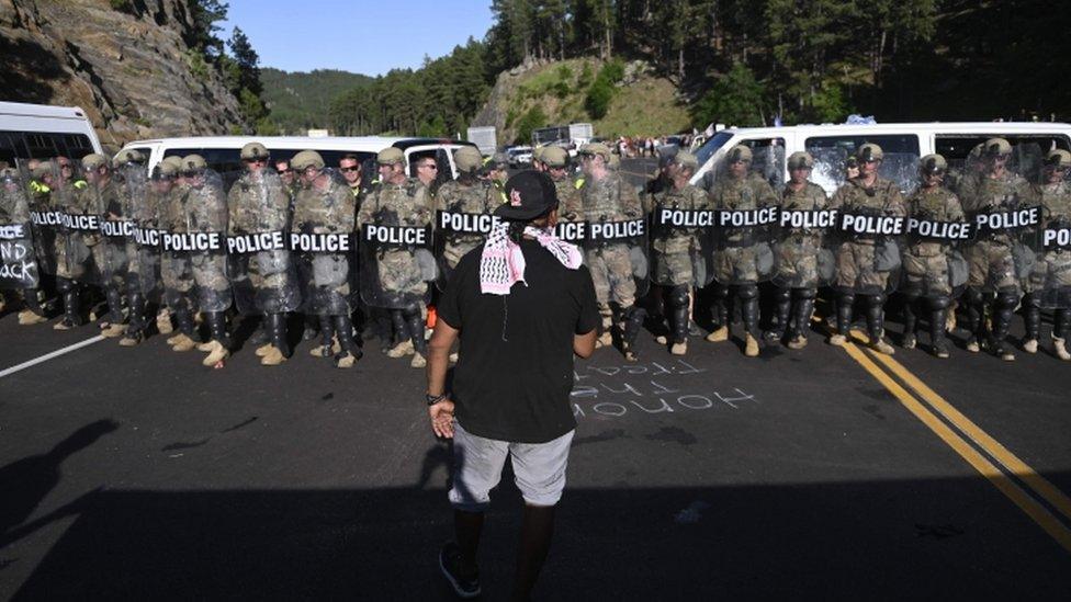 A man faces a row of police as activists and members of different tribes from the region block the road to Mount Rushmore National Monument in Keystone, South Dakota