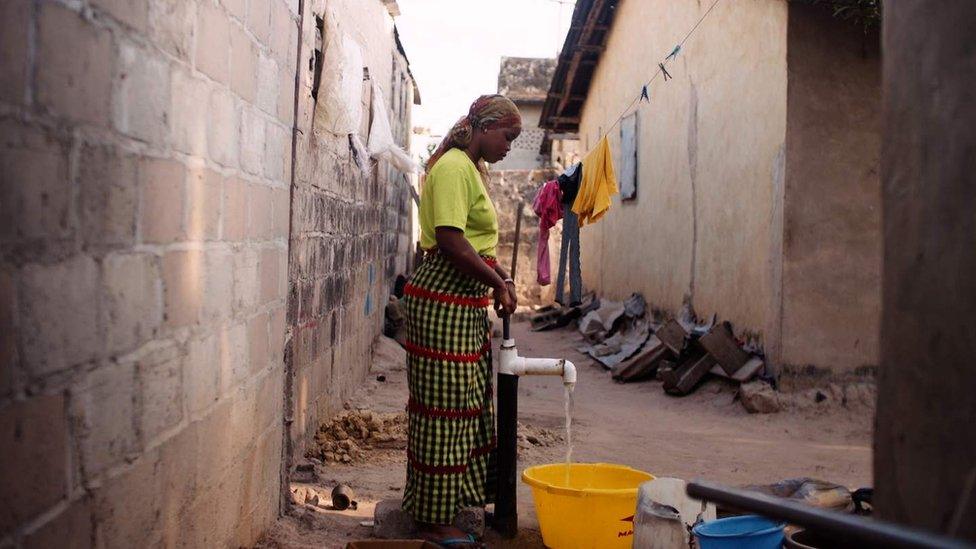 A woman standing in an alleyway pumping water from a borehole into a bucket.