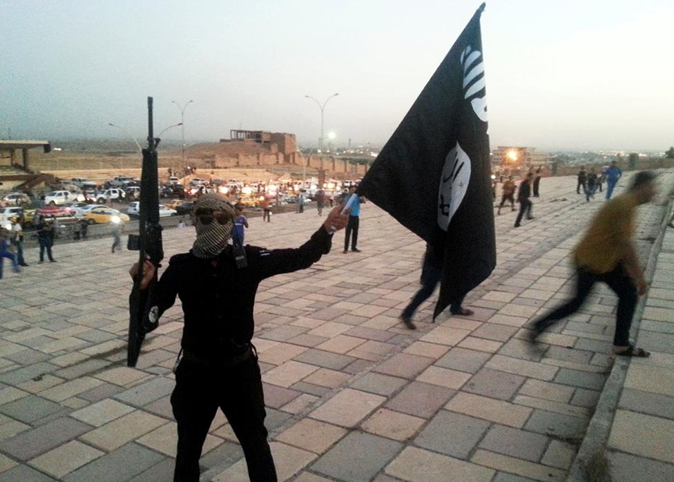 A fighter of the Islamic State of Iraq and the Levant (ISIL) holds an ISIL flag and a weapon on a street in the city of Mosul, June 23, 2014.