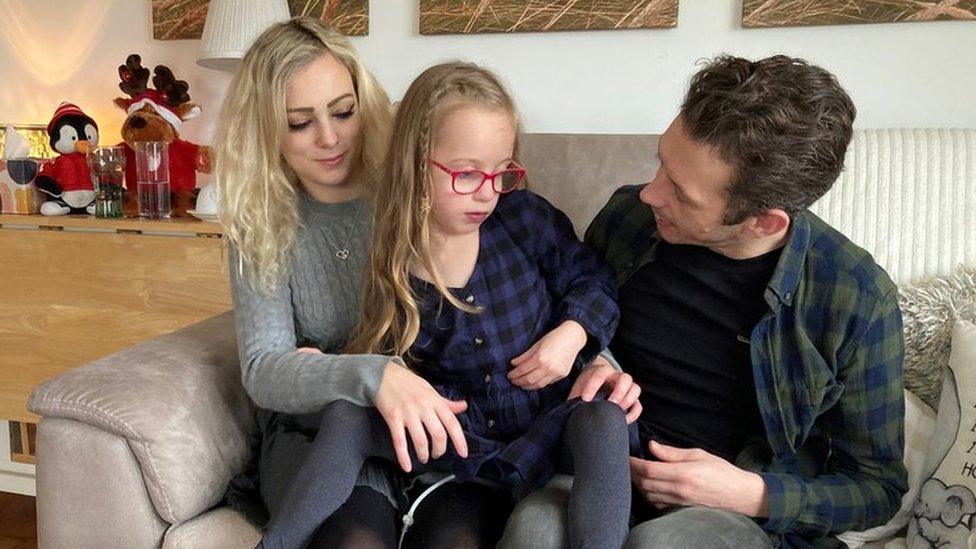 Minnie, with long blonde hair and red glasses, sits between her mother Charlotte and father Jesse