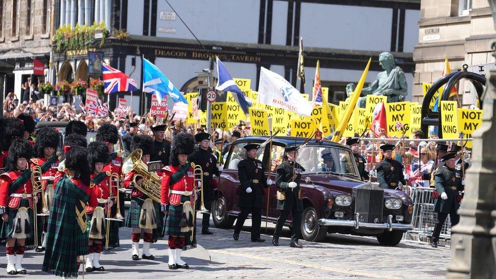 Car carrying the King and Queen enter the square at St Giles, one side of the road has Union flags and Saltire flags, the other has protesters waving yellow 'not my king' placards