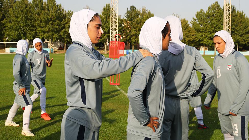 Iran Women's National Football during a training session. There are six women in the pictures, wearing grey tracksuits and a white hijab. The woman in the centre of the picture has her right hand on the shoulder of the woman in front. They are standing on a green pitch with trees visible in the background and a white metal structure.