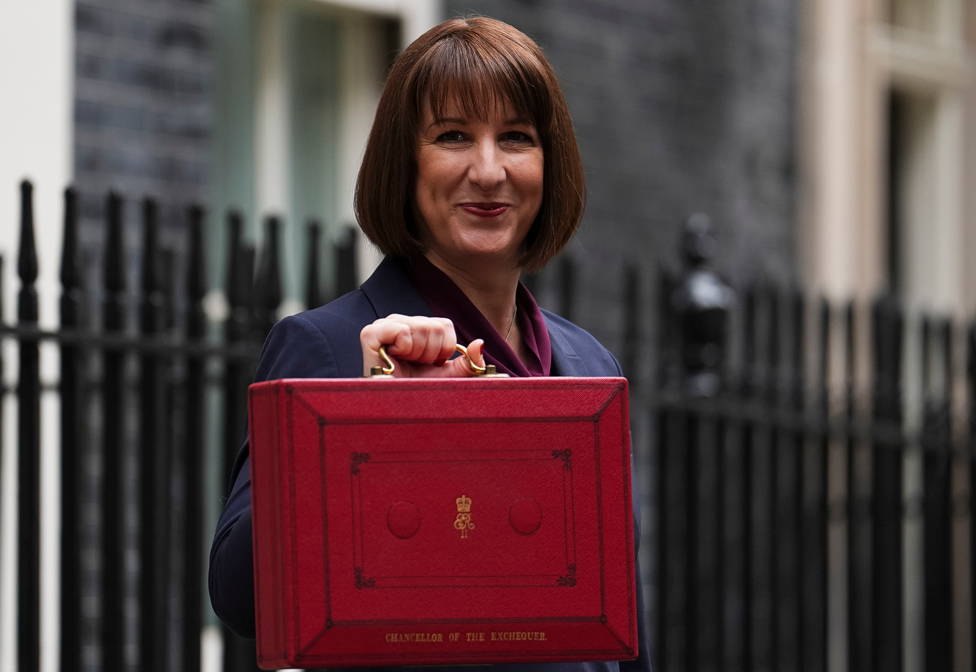 Chancellor Rachel Reeves holds up her red budget box outside the black door to number 11 Downing Street on Wednesday morning, before heading to the House of Commons to make her statement. The box has the words "Chancellor of the Exchequer" printed on it in gold lettering.