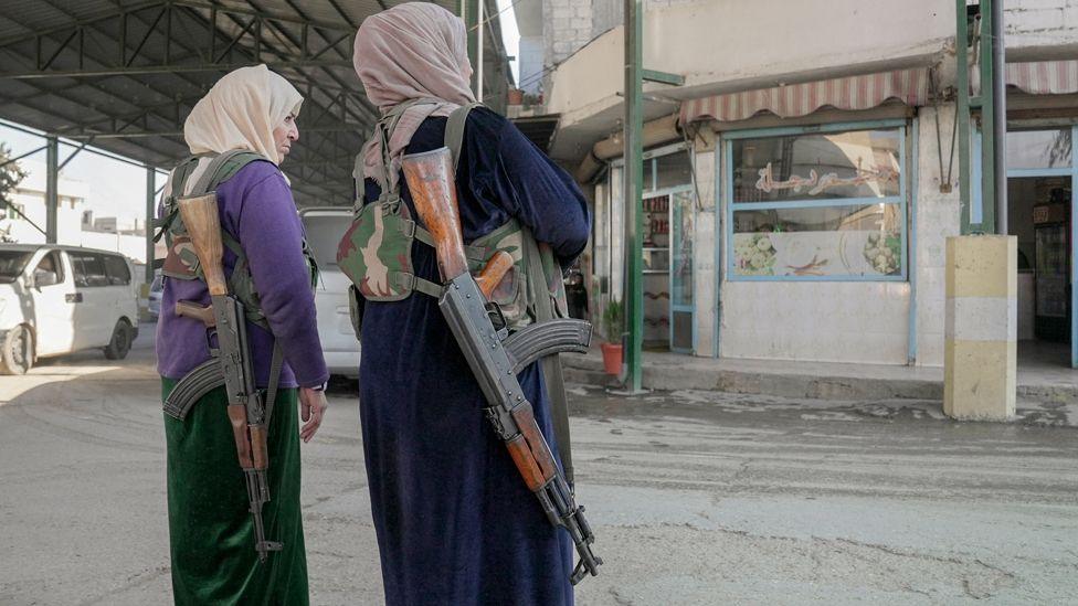 Two women in with AK-47 assault rifles slung over their shoulders. They are wearing long dresses and headscarves.