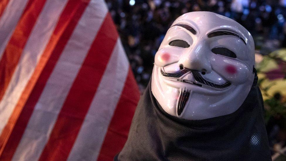 An Anonymous mask is seen next to a US flag in this photograph, which was actually taken in Hong Kong during the pro-democracy protest there in 2019