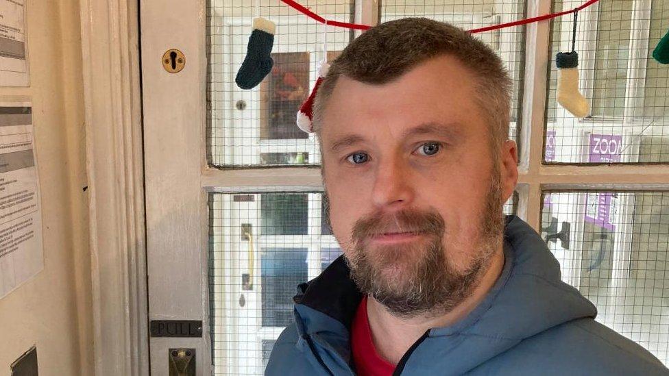 Man with short dark hair and beard standing by a door