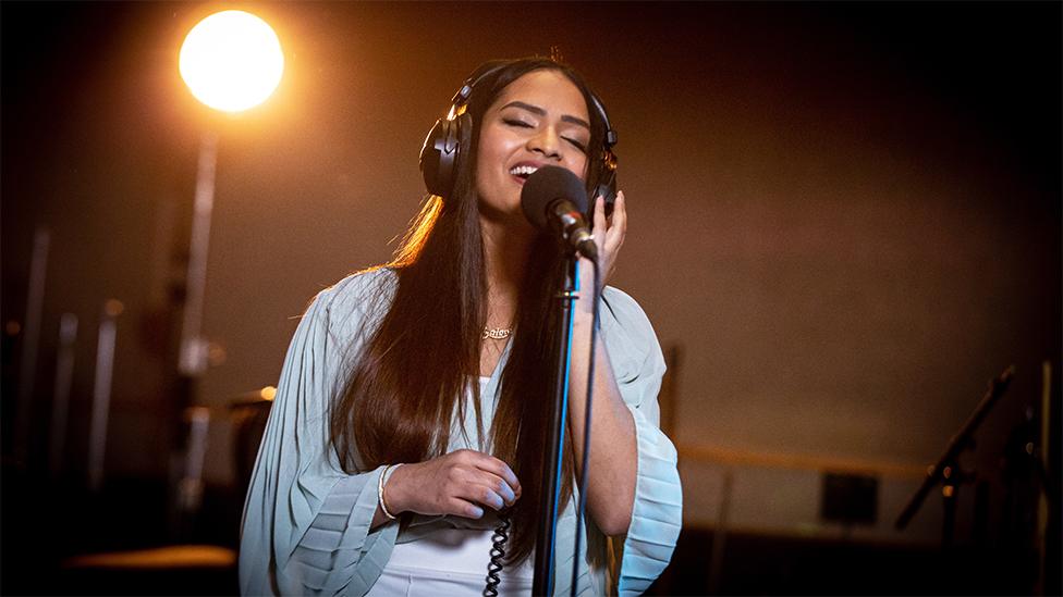 Saloni, an Asian woman, closing her eyes and singing into a black microphone while wearing black headphones. Behind her is black background and a spotlight shining on her. The top part of her outfit is light blue.