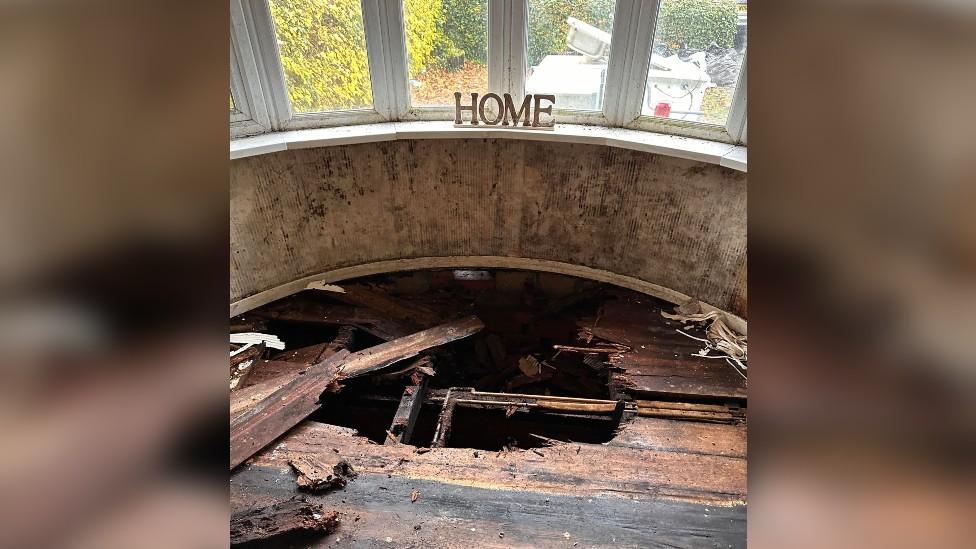 A decoration spelling "home" on a mouldy windowsill above rotten floorboards