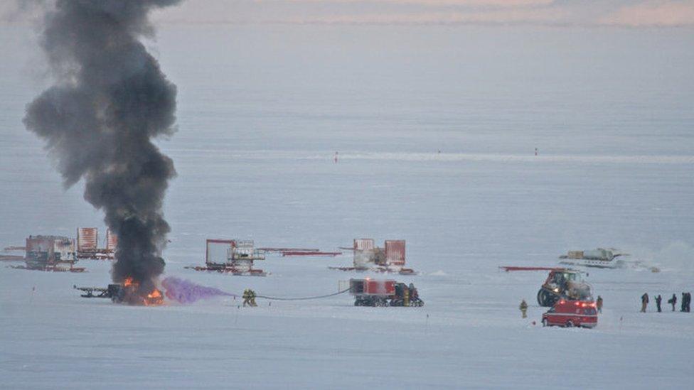 Smoke rises from a vehicle fire on an ice field. Fire-fighters spray a purple chemical on it