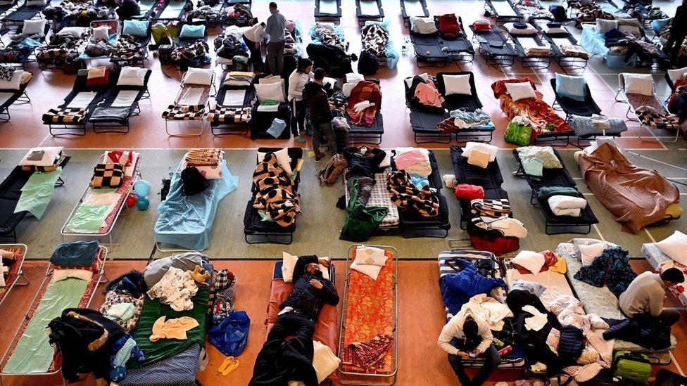 Aerial view of dozens of camp beds and people in sports hall