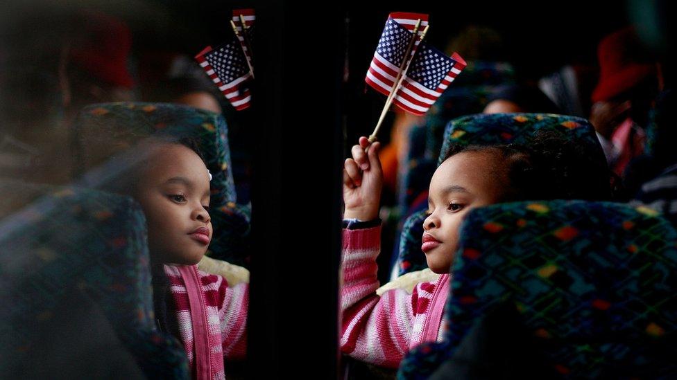 A small black girl waves an American flag, while looking out of the window of a train