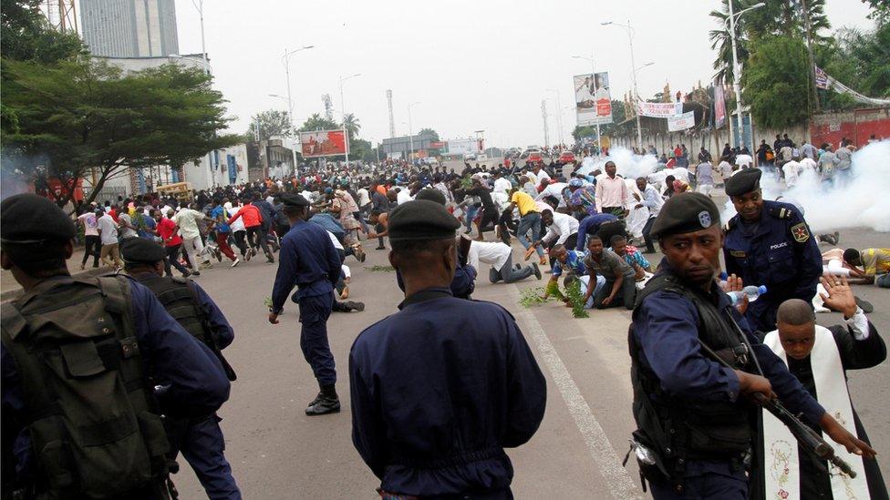 Riot policemen fire teargas canisters to disperse demonstrators during a protest - many people, including a priest, can be seen kneeling, hands raised