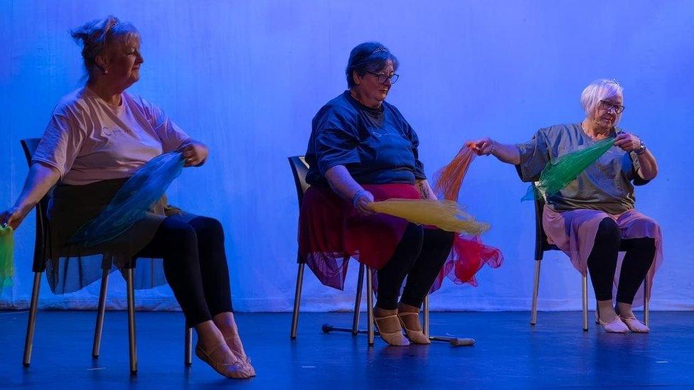 Three women sit on chairs on a stage and rhythmically move coloured handkerchiefs around