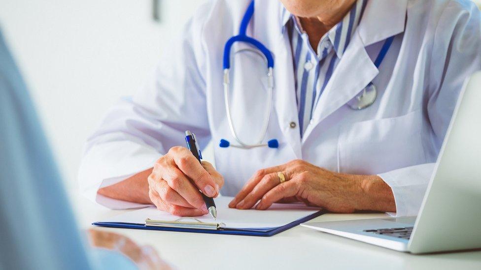 The arms and torso of a doctor at their desk, wearing a white coat and writing notes