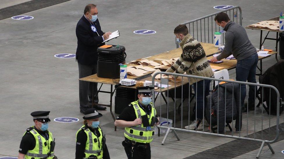 Alba party leader Alex Salmond watches staff about to start counting, while three police officers walk past