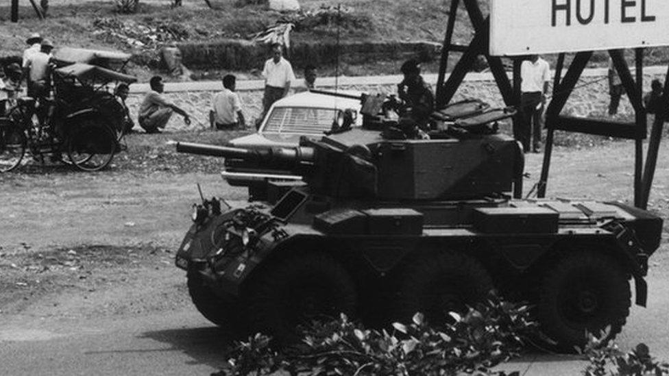 Young people stood outside an hotel in Indonesia with a military armoured vehicle on stand by