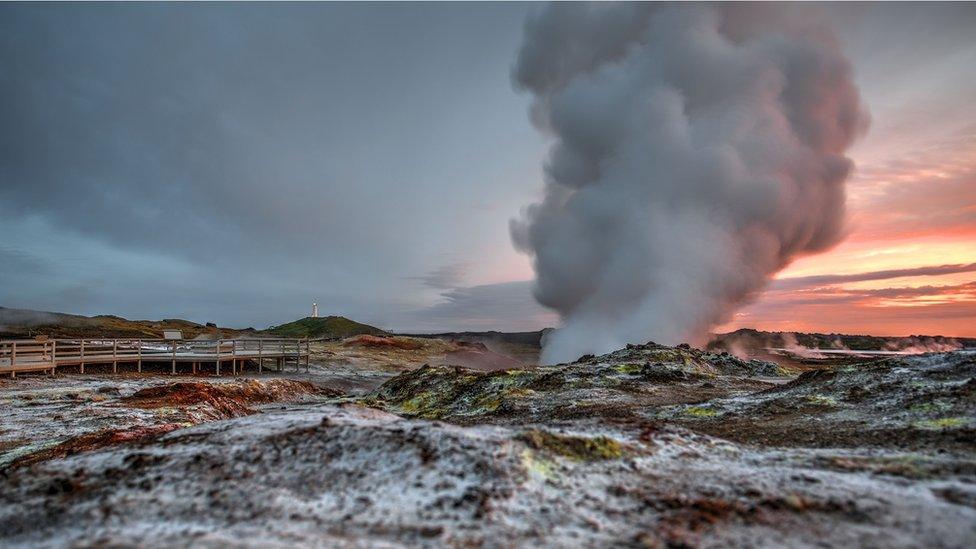 Steam emerges from a geothermal vent, a sunset in the background