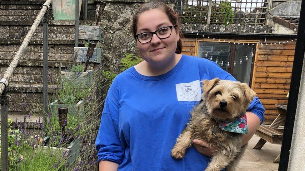 Young white woman wearing glasses holding a dog under her arm in a garden with unlit fairylights behind her