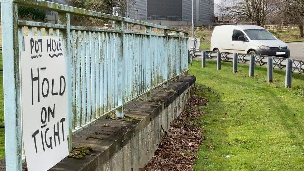 "Hold on tight" sign on blue railings in the middle of a roundabout. A white van is passing.