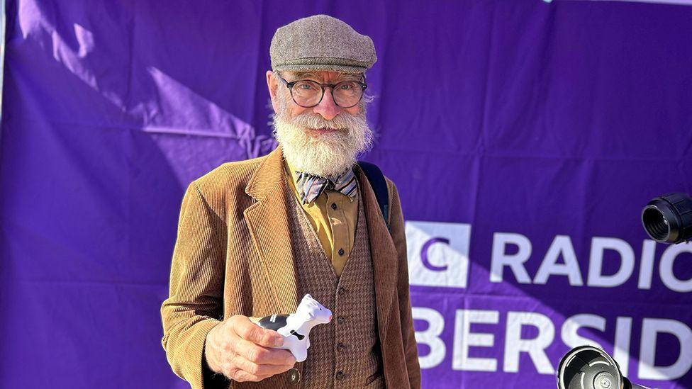 Rob Bator, dressed in brown tweeds with a bow tie, a grey flat cap, glasses and a large white beard, holds a small rubber black and white stress cow at the Driffield Show