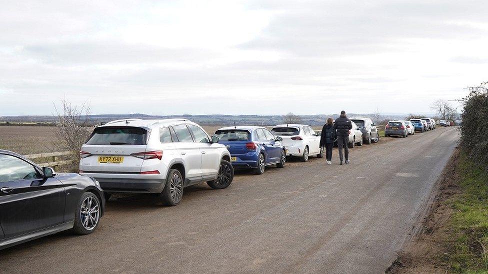 Cars parked on verge as pedestrians walk along road