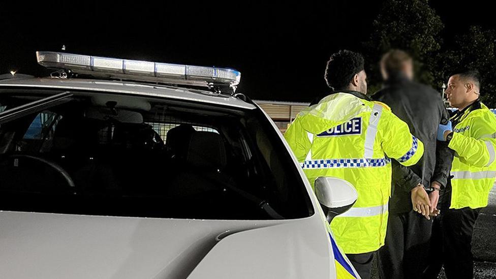 Officers with a man in handcuffs next to a police car