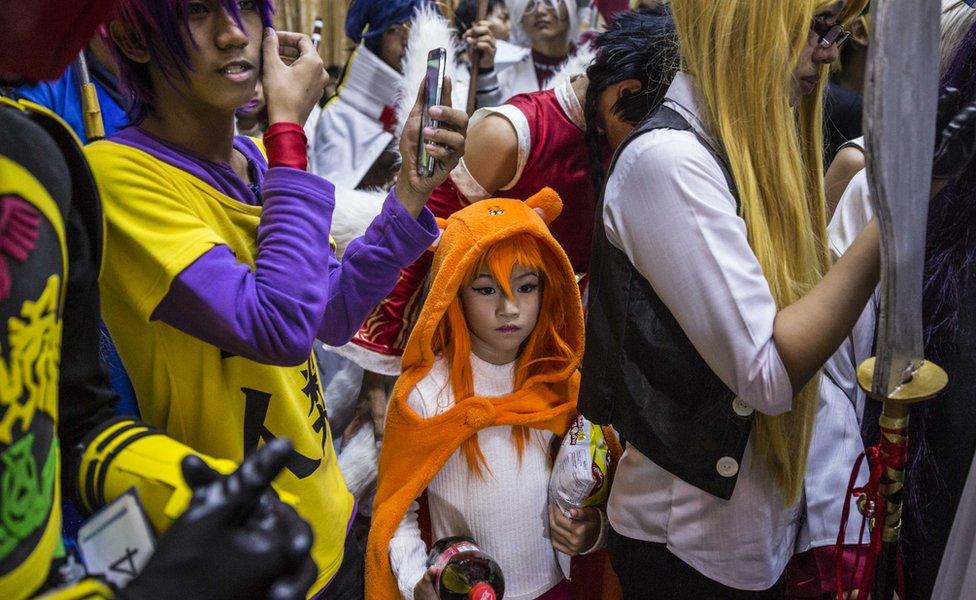 A girl in an orange animal outfit surrounded by taller, older boys in colourful wigs