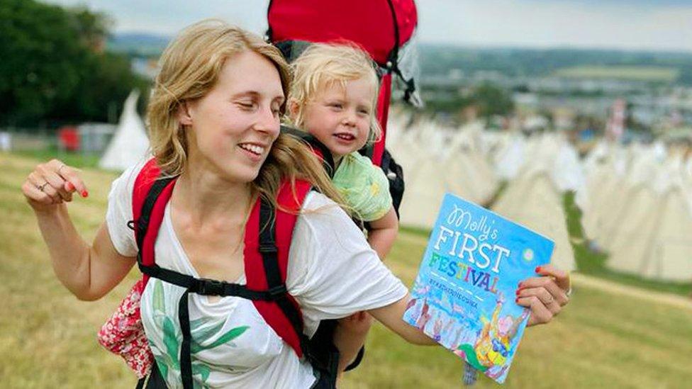 A young blonde haired woman with a young toddler in a baby carrier on her back