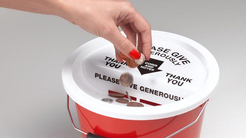 A hand drops coins into a red charity bucket