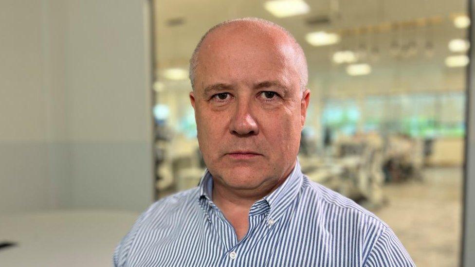 Man with very short hair wearing a blue and white striped shirt and standing in a large office