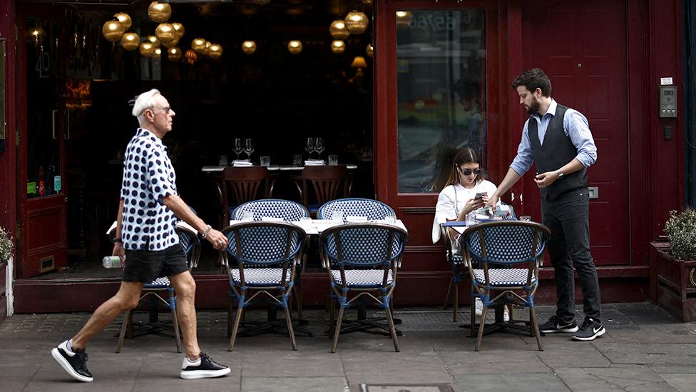 A customer is served outside an Italian restaurant, with a pedestrian walking in the foreground, in the Soho area of London on June 18, 2023