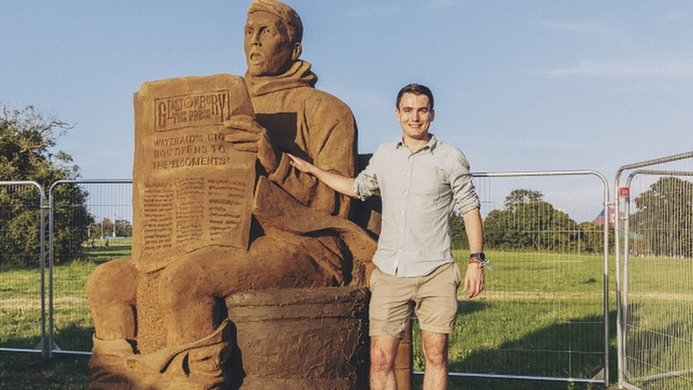 A mud and sand sculpture of a man on a toilet, reading a newspaper with the man standing next to it