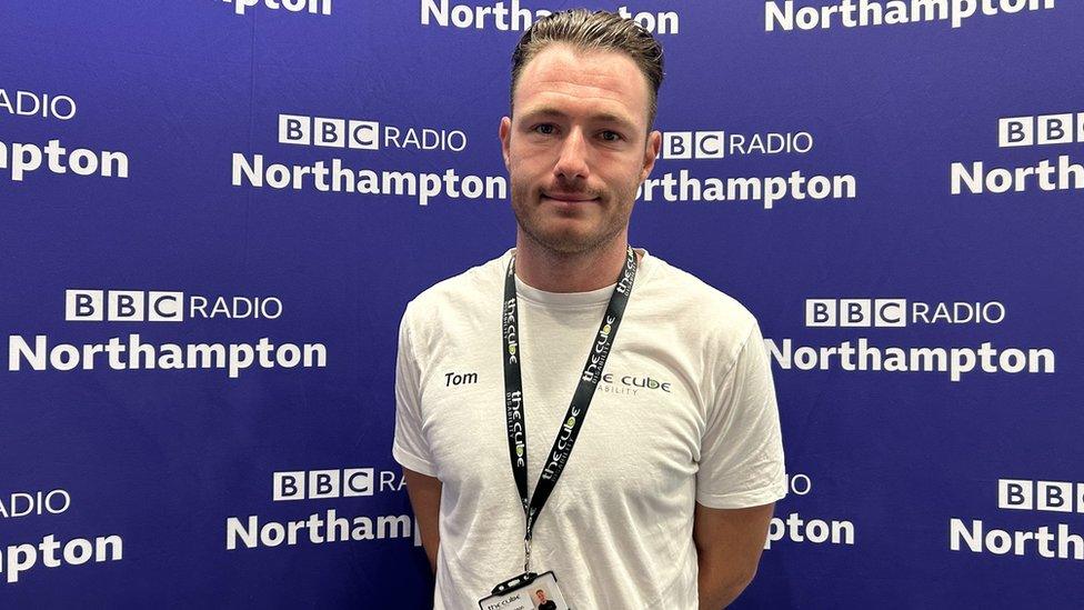 Man with short hair wearing white T-shirt and lanyard in front of BBC Radio Northampton branding